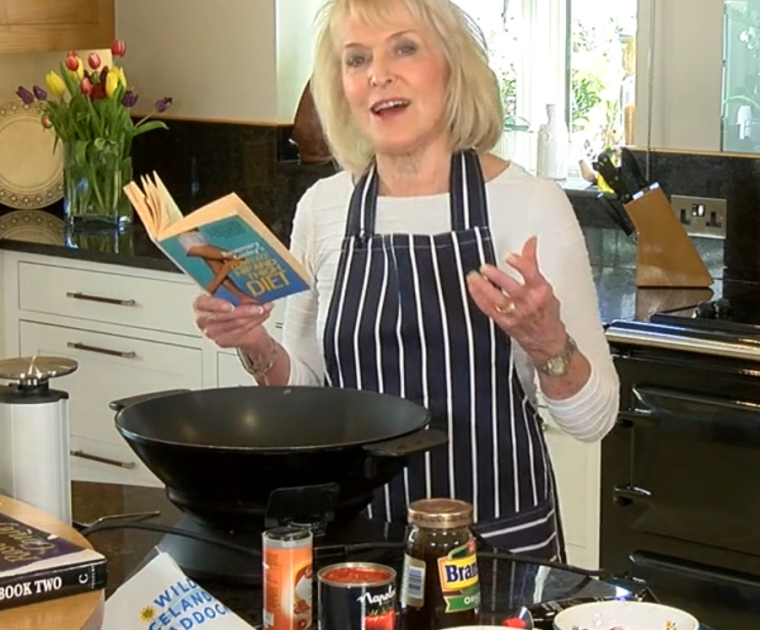 Rosemary Conley in the kitchen with an electric wok reading a recipe for Fish Curry out of her Hip & Thigh Diet book