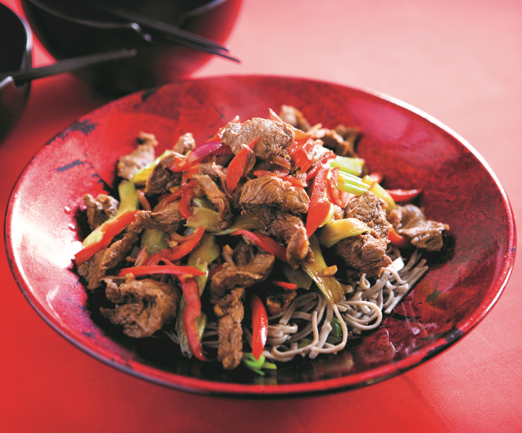 A stirfry of Beef steak strips with ginger, sugar-snap peas, and pak choi on a bed of nnodles on a red oriental style bowl