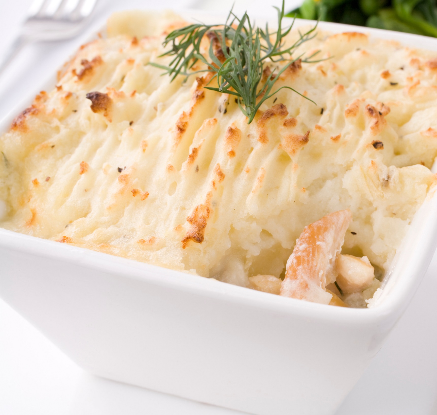 A square white bowl containing smoked haddock in a creamy sauce, with a mashed potato topping garnished with dill.