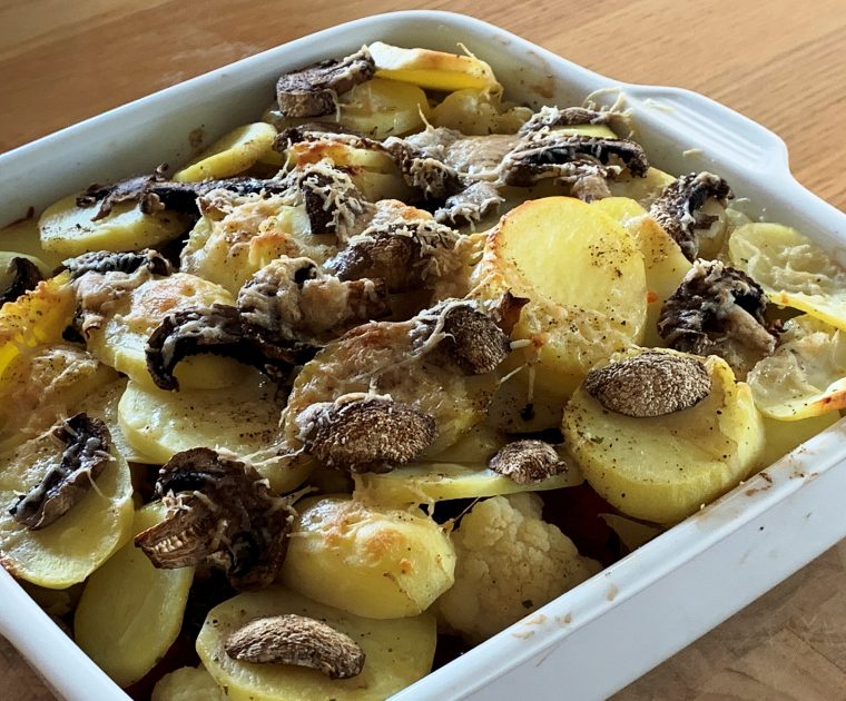A square white dish with layers of mushroom on top of slices of potato sprinkled with parmesan cheese. Below is a bake of vegetables and baked beans.