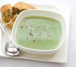 A white bowl of Leek and Broccoli soup with a granary roll and soup spoon beside.