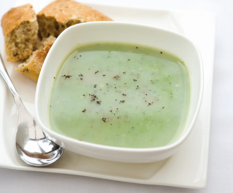 A white bowl of Leek and Broccoli soup with a granary roll and soup spoon beside.