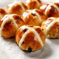 A set of glistening hot cross buns with a shiny glaze, a rich brown colour and some currants peeking out.
