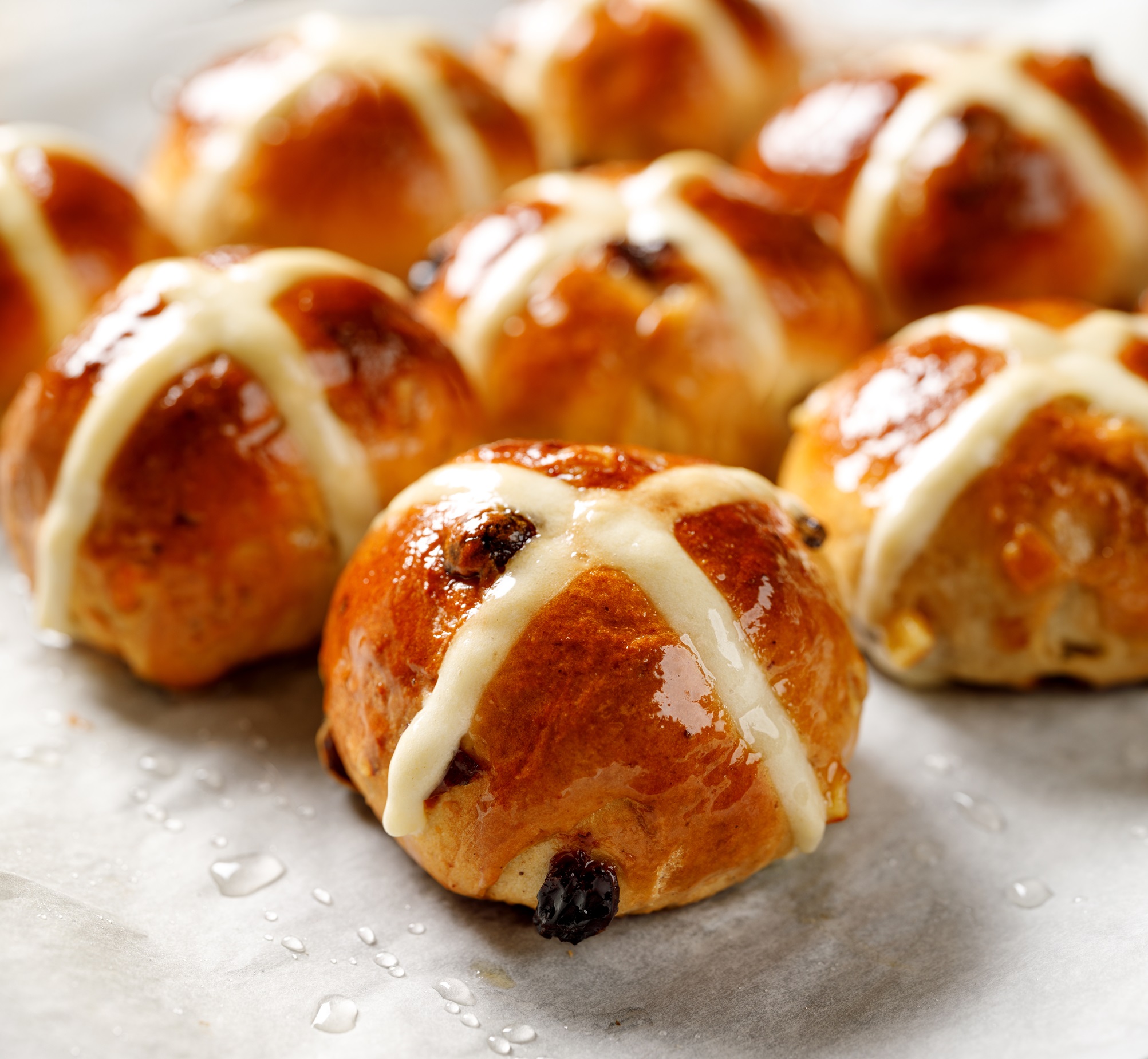 A set of glistening hot cross buns with a shiny glaze, a rich brown colour and some currants peeking out.