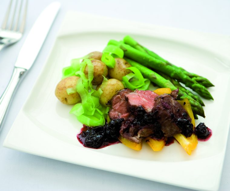 Lamb Medallions with a blackcurrant sauce served on a white plate with new potatoes with their skins on, chopped leeks and asparagus tips.