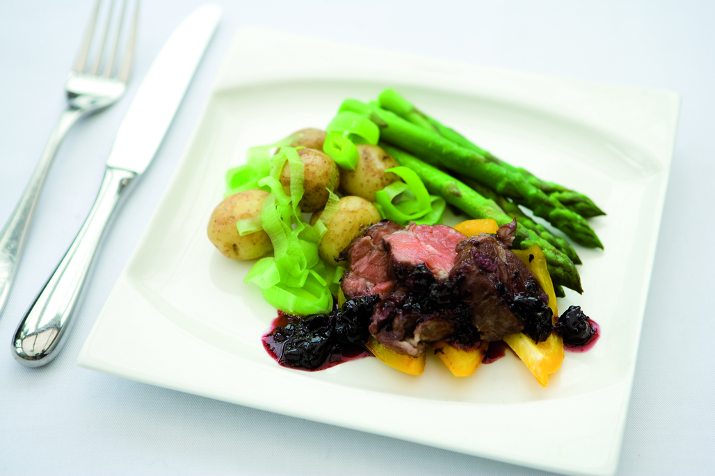 Lamb Medallions with a blackcurrant sauce served on a white plate with new potatoes with their skins on, chopped leeks and asparagus tips.