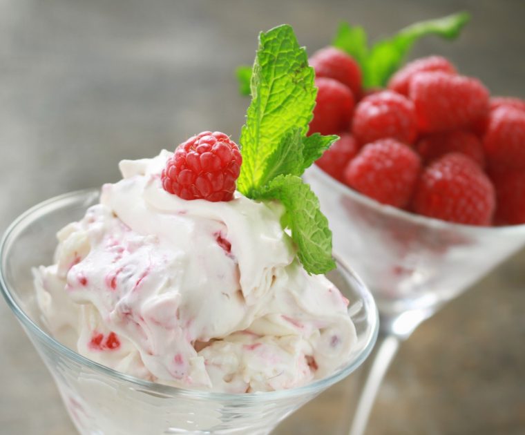 Two martini glasses - one containing Eton Mess made from greek yogurt, raspberries and meringue garnished with a sprig of mint, the other containing further fresh raspberries.