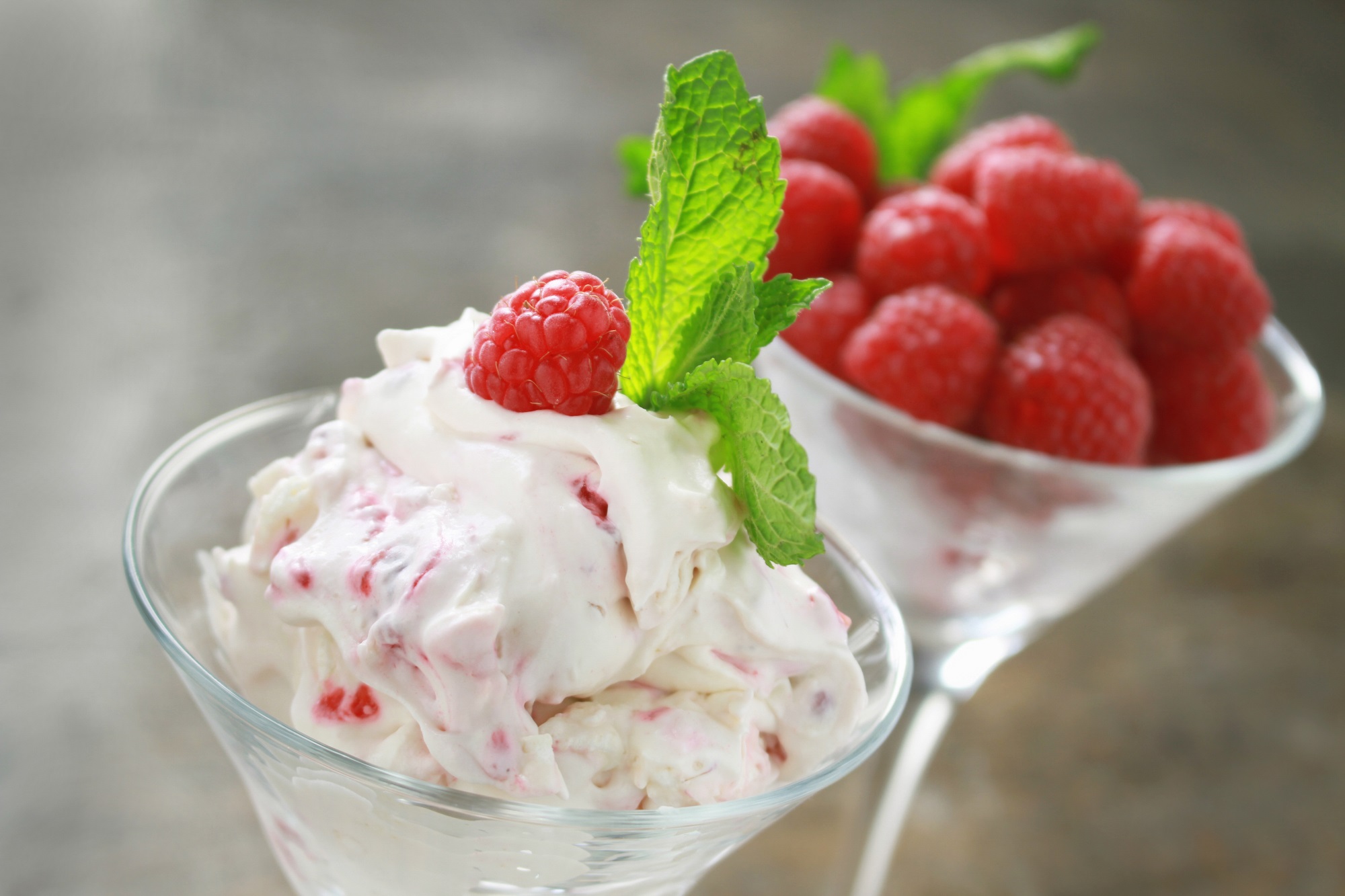 Two martini glasses - one containing Eton Mess made from greek yogurt, raspberries and meringue garnished with a sprig of mint, the other containing further fresh raspberries.