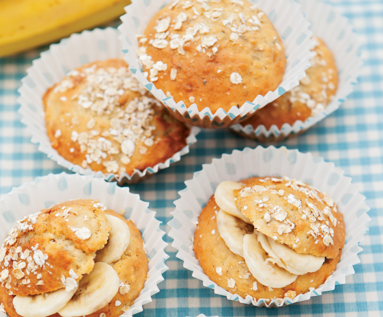 Banana Muffins in paper cases. Some are decorated by being sprinkled with oats, others have a slit containing slices of banana in the top.