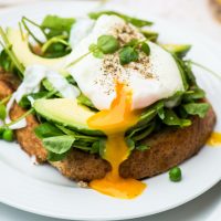 Poached Egg & Avocado on Toast - A poached egg with the yolk oozing out of it sits on top of slices of avocado on a bed of watercress and baby spinach on a slice of wholemeal toast.