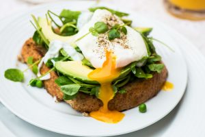 Poached Egg & Avocado on Toast - A poached egg with the yolk oozing out of it sits on top of slices of avocado on a bed of watercress and baby spinach on a slice of wholemeal toast.