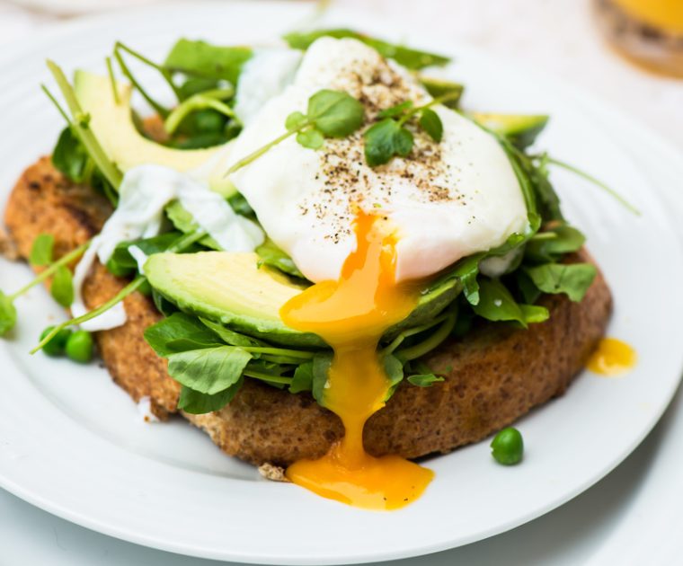 Poached Egg & Avocado on Toast - A poached egg with the yolk oozing out of it sits on top of slices of avocado on a bed of watercress and baby spinach on a slice of wholemeal toast.