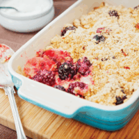 A rectangular oven-proof dish co0ntaining apple and blackberry spelt crumble - a fruit mix with a spelt-flour crumble topping. One spoonful has been taken out so you can see the delicious fruit filling under the crumble topping.