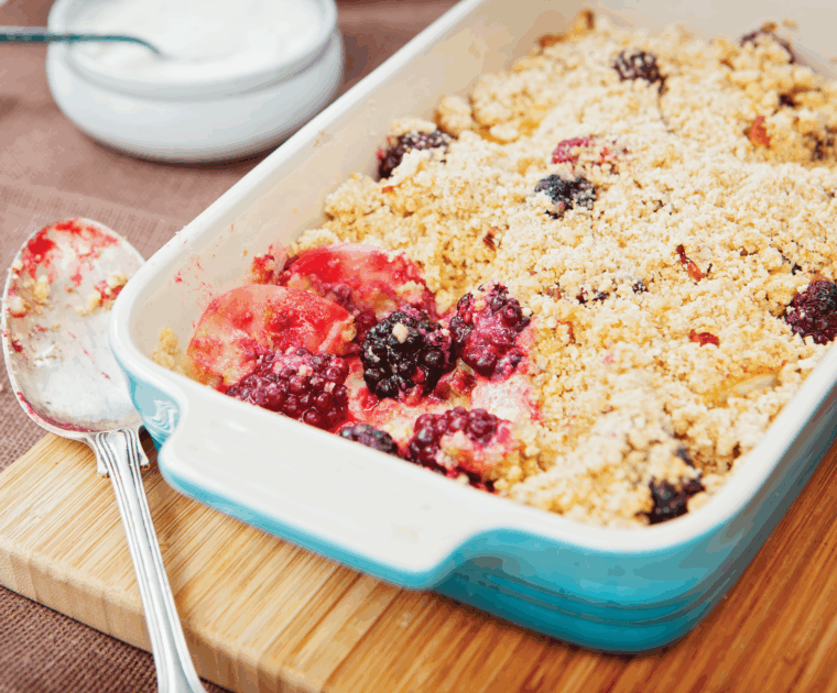 A rectangular oven-proof dish co0ntaining apple and blackberry spelt crumble - a fruit mix with a spelt-flour crumble topping. One spoonful has been taken out so you can see the delicious fruit filling under the crumble topping.