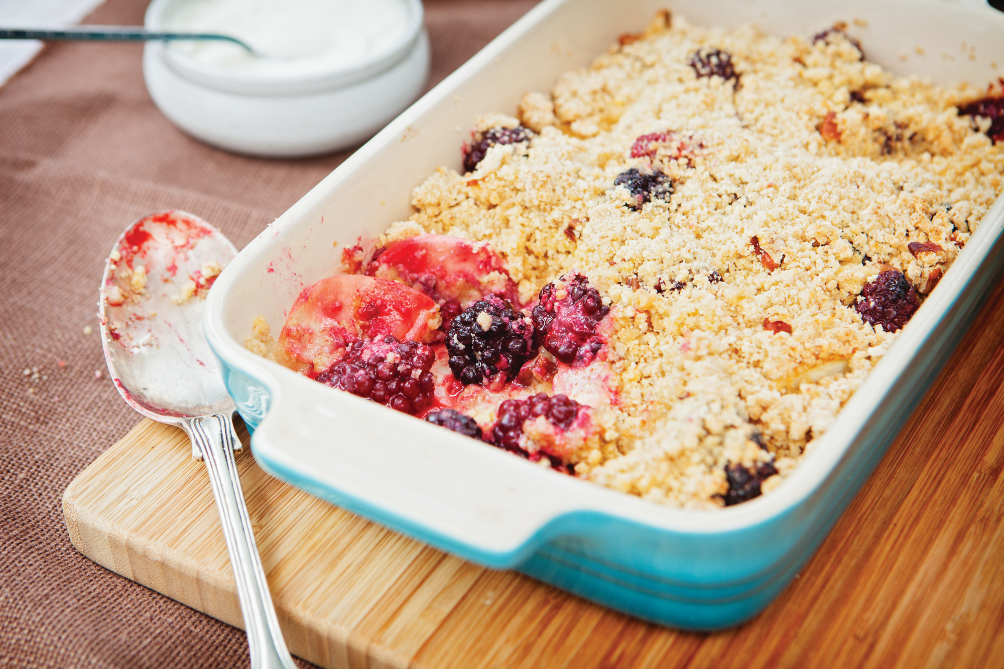 A rectangular oven-proof dish co0ntaining apple and blackberry spelt crumble - a fruit mix with a spelt-flour crumble topping. One spoonful has been taken out so you can see the delicious fruit filling under the crumble topping.