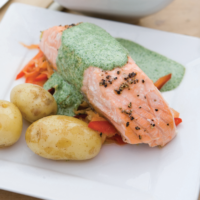 A baked salmon steak drizzled with watercress sauce on a bed of assorted mixed vegetables served with some boiled new potatoes.