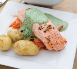 A baked salmon steak drizzled with watercress sauce on a bed of assorted mixed vegetables served with some boiled new potatoes.