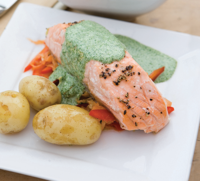 A baked salmon steak drizzled with watercress sauce on a bed of assorted mixed vegetables served with some boiled new potatoes.