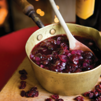 Cranberry and Red Wine Sauce being stirred with a wooden spoon in a saucepan on a chopping board surrounded with cranberries.