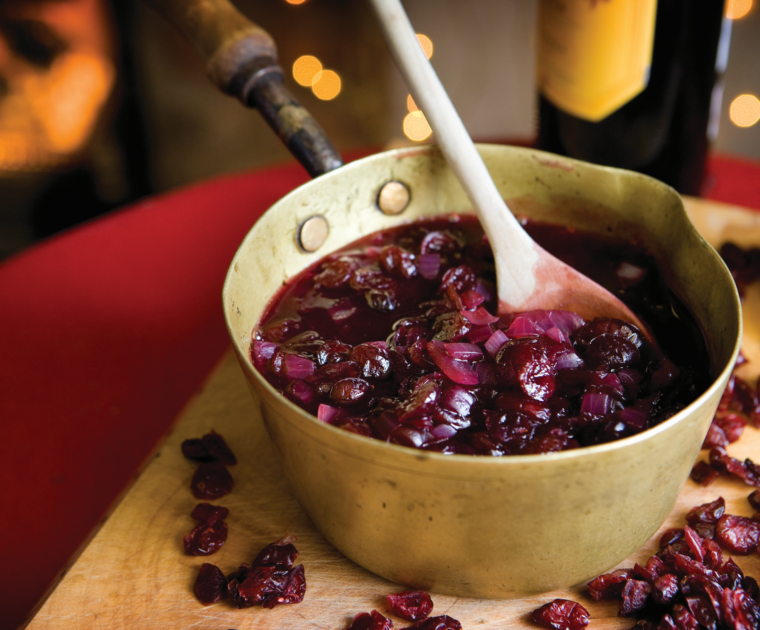 Cranberry and Red Wine Sauce being stirred with a wooden spoon in a saucepan on a chopping board surrounded with cranberries.