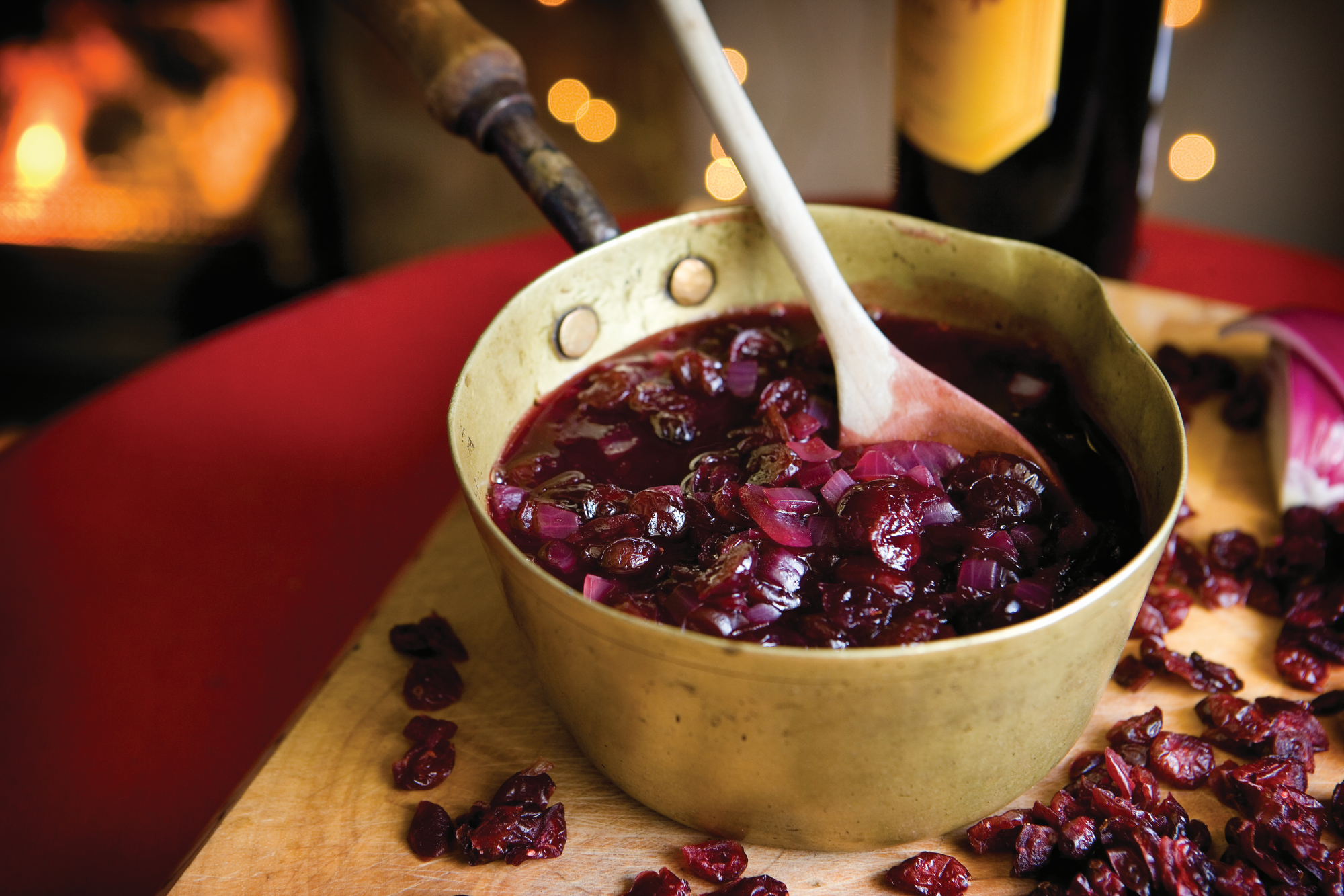 Cranberry and Red Wine Sauce being stirred with a wooden spoon in a saucepan on a chopping board surrounded with cranberries.