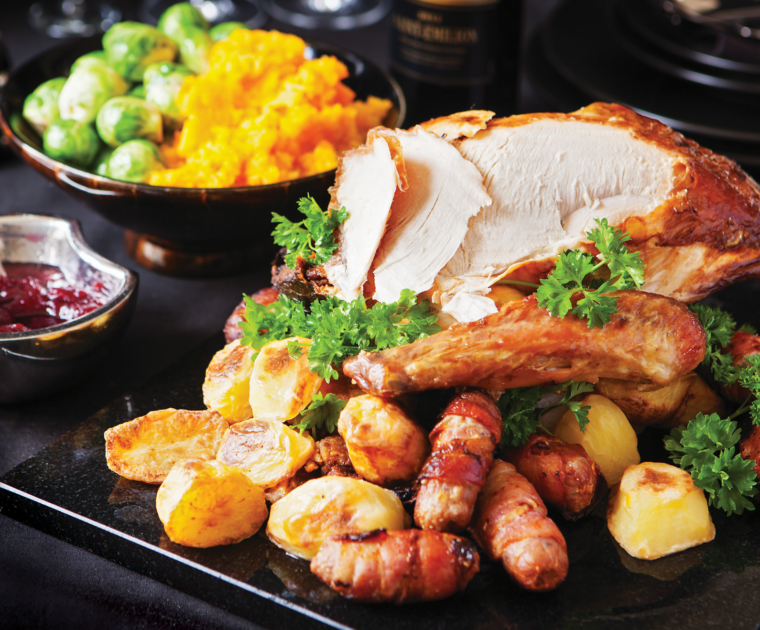 A roast turkey crown sitting on a serving board with a few slices already cut. It is surrounded by roast potatoes and pigs in blankets garnished with parsley. On the side is a bowl of Brussells sprouts and swede and a dish of cranberrry sauce