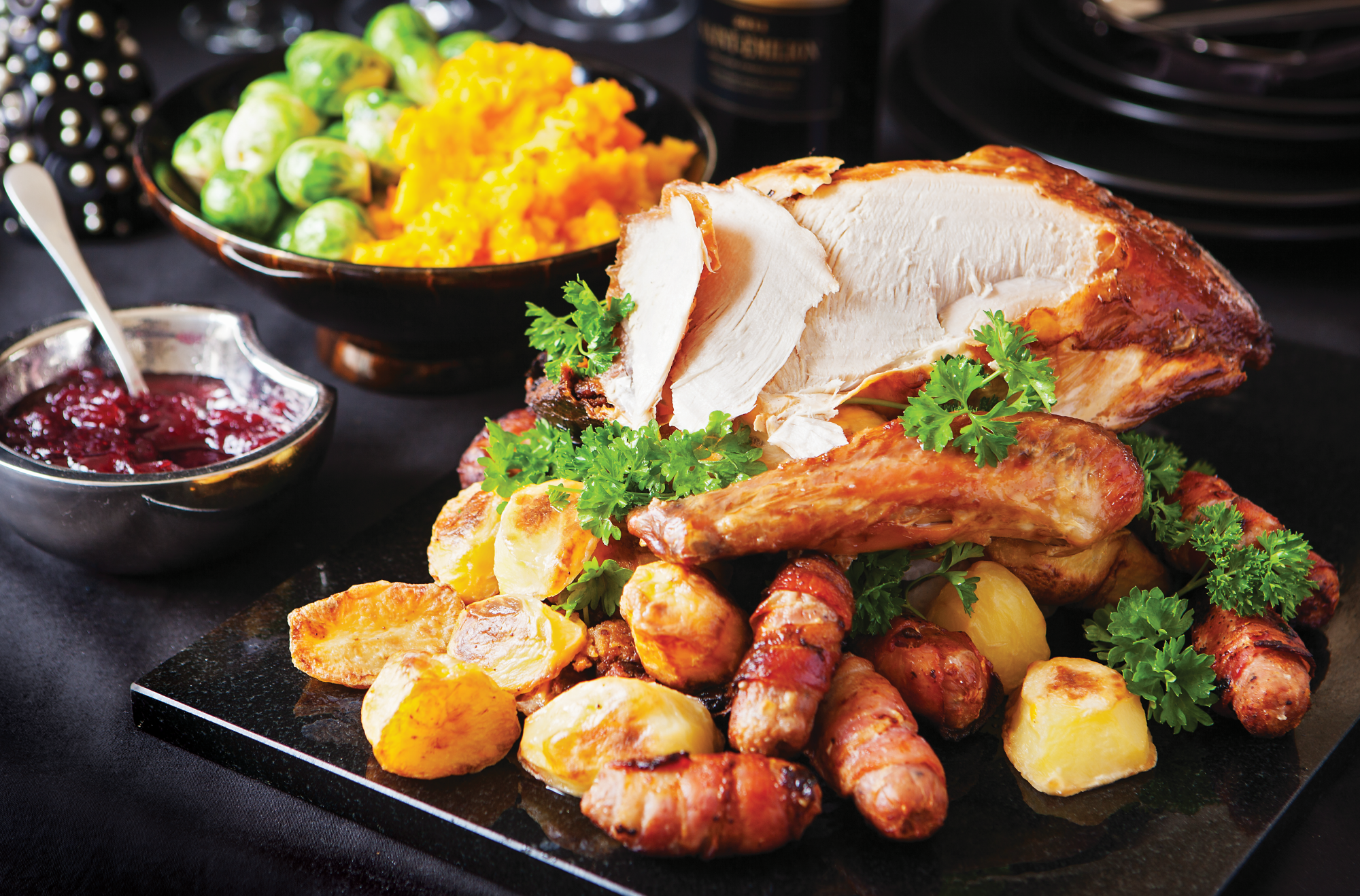 A roast turkey crown sitting on a serving board with a few slices already cut. It is surrounded by roast potatoes and pigs in blankets garnished with parsley. On the side is a bowl of Brussells sprouts and swede and a dish of cranberrry sauce