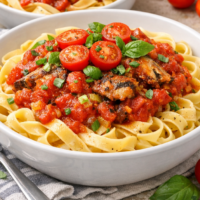 A bowl full of tagliatelle past topped with a sauce made with sardines and tinned tomatoes with garlic and leek and garnished with fresh cherry tomatoes and basil.